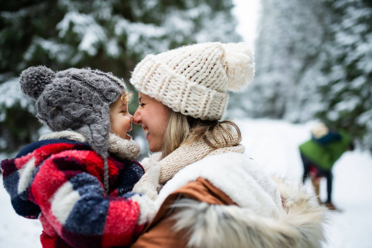 Amidst snow-covered trees, a mother and child in cozy winter attire and knitted hats share a tender moment with their noses touching in the snowy forest. This serene scene captures the warmth of togetherness.