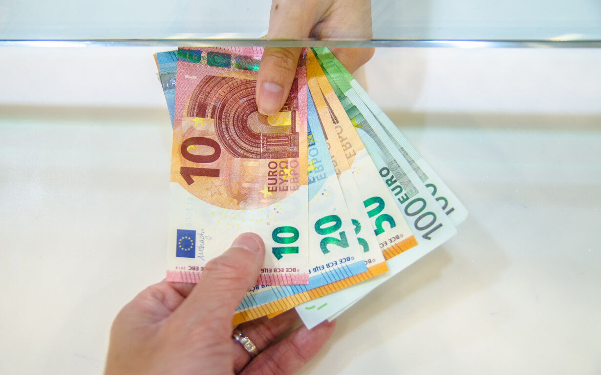 Close-up of a person handing Euro banknotes through a transparent partition. The denominations include 10, 20, and 50 Euro notes. The recipient's hand is visible on the other side, receiving the money. The scene suggests a financial transaction or exchange, possibly to send remittances.