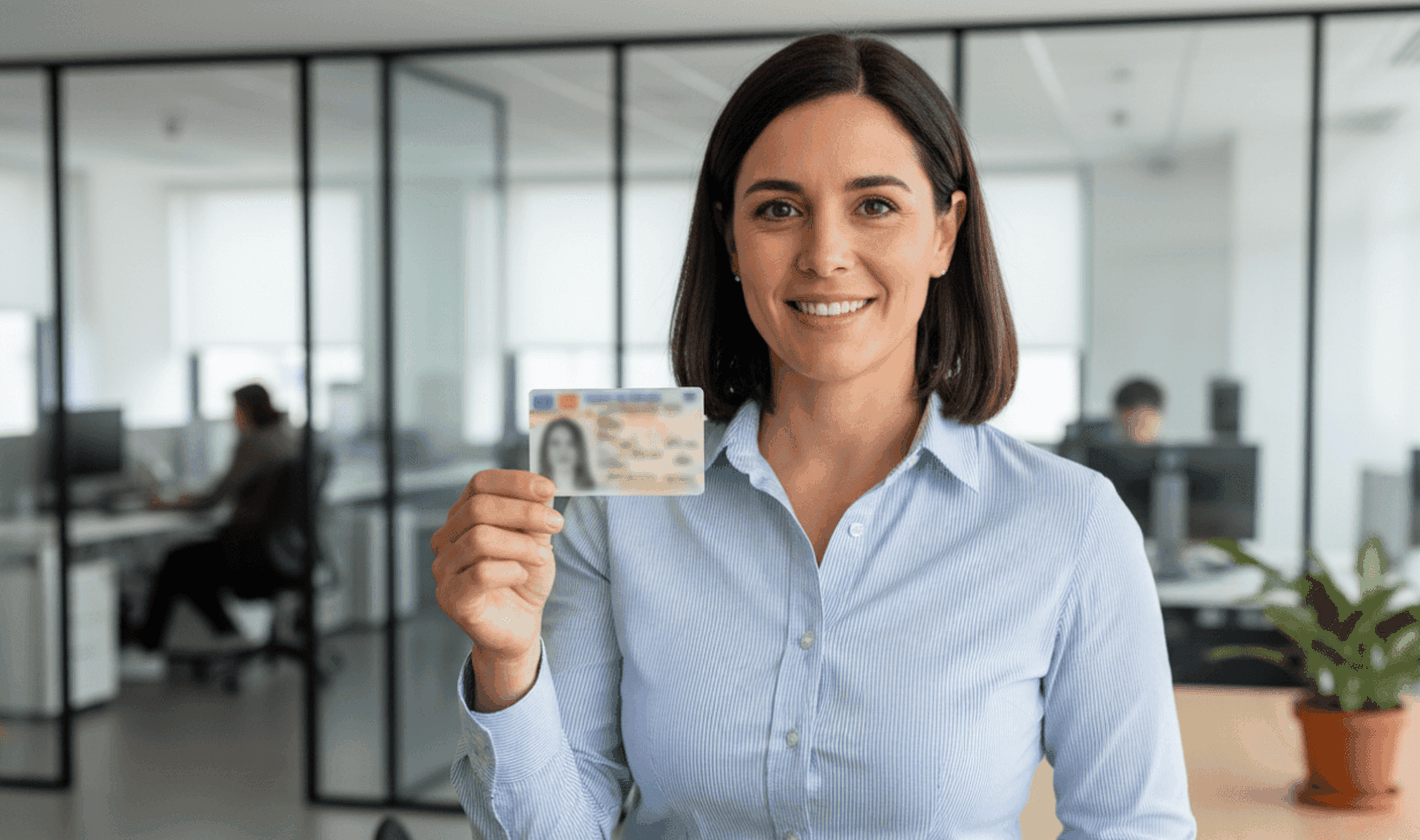A smiling woman in a light blue shirt stands in an office, holding up an ID card to verify your identity. The background features glass partition walls, office furniture, and people working at desks.