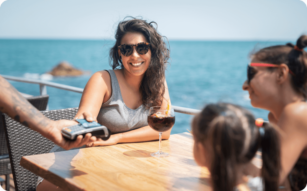 Smiling woman wearing sunglasses sits at a wooden table by the sea, holding a drink with a slice of orange. Two others are at the table, including a child. A person hands over a credit card payment terminal. Sunny and relaxed beachside setting.