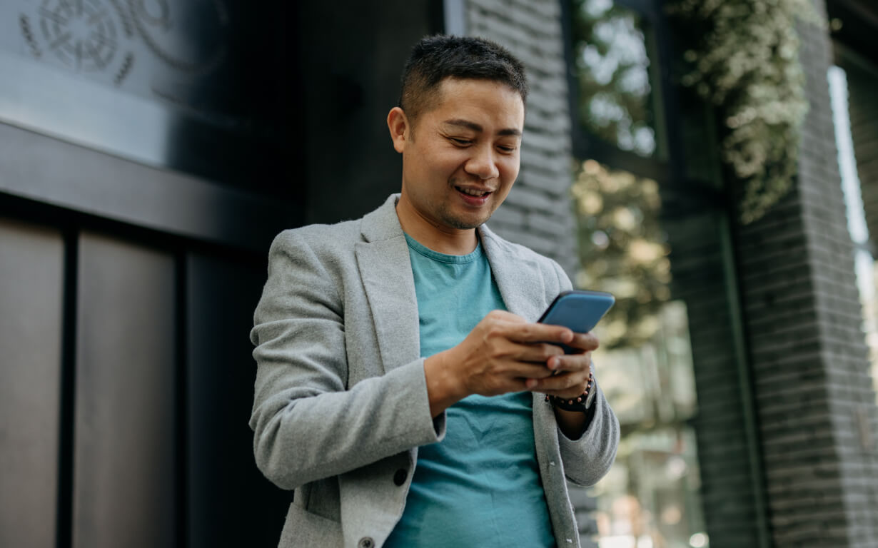 A person in a gray blazer and teal shirt is smiling down at their smartphone, while standing outdoors in front of a modern building with brick walls.
