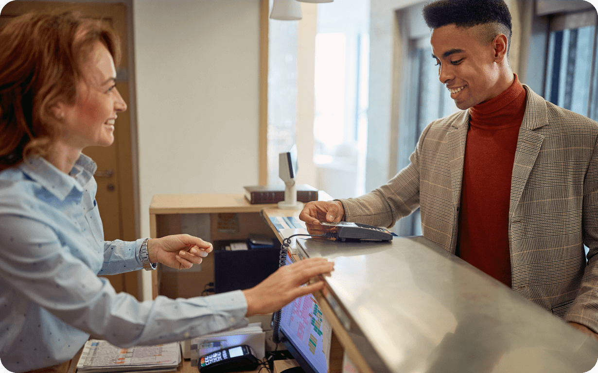A smiling man in a suit places his card on a payment terminal at a reception desk, opposite a friendly receptionist in a light blue blouse. The setting appears modern and well-lit, with the transaction creating a welcoming atmosphere.