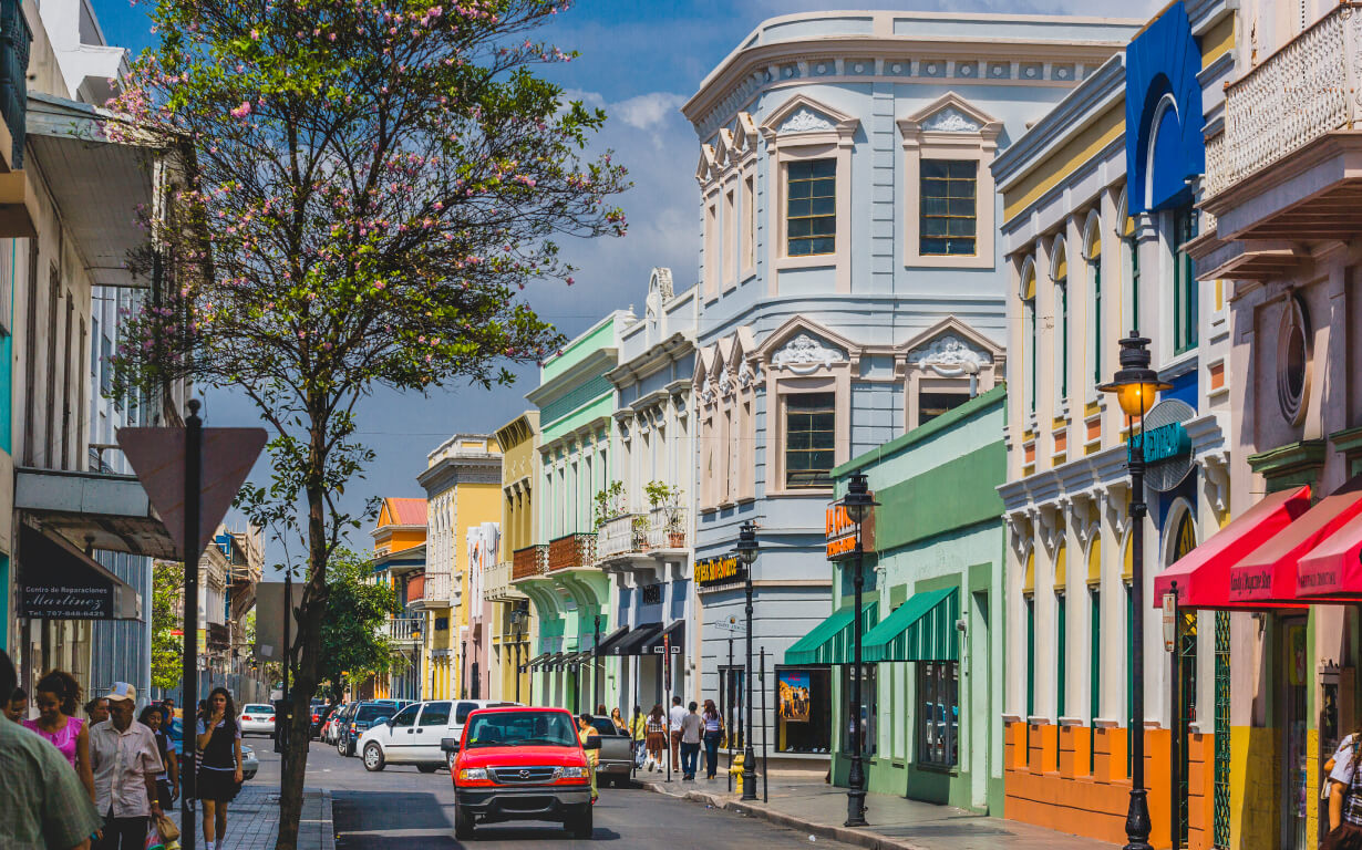 A vibrant street scene in Puerto Rico features colorful colonial-style buildings, with pastel facades in blue, green, yellow, and pink. People stroll along the sidewalk as a red and white vehicle drives by. A tree with pink blossoms stands on the left—an idyllic setting for those moving to Puerto Rico.