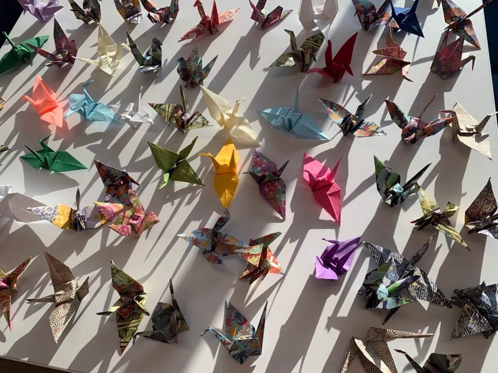 A display of colorful origami paper cranes arranged on a white surface, with sunlight casting dramatic shadows behind them.