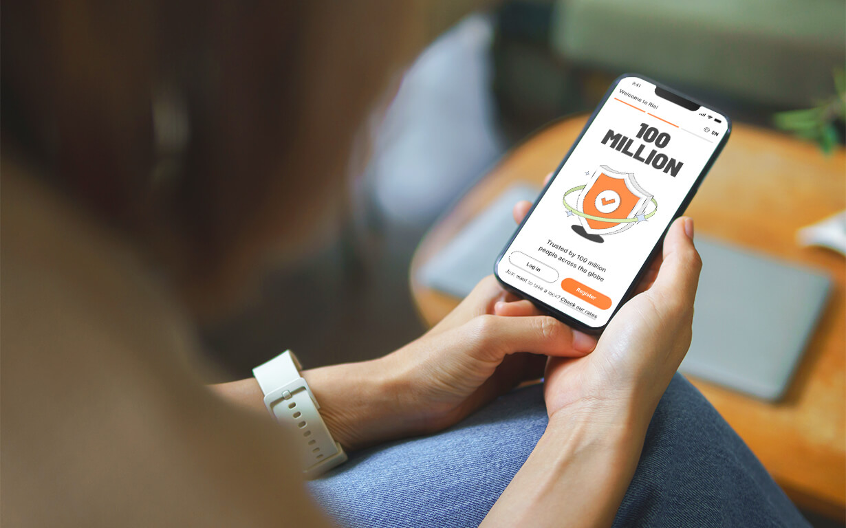 A person wearing a smartwatch holds a smartphone displaying the Ria app screen that says “100 MILLION” above a shield with a heart, and buttons labeled “Get Info” and “Donate Now.”.