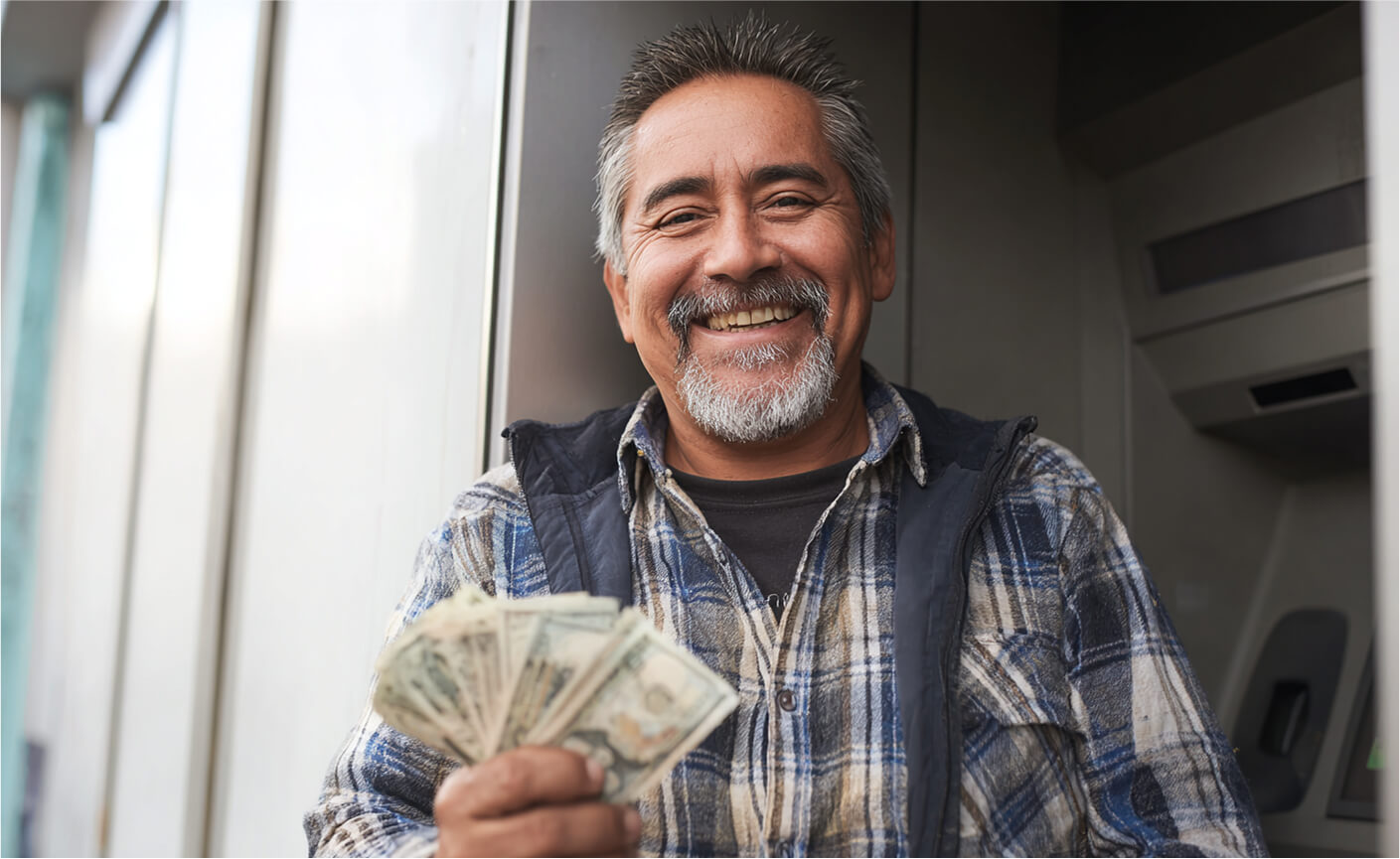 Smiling man in a plaid shirt stands in front of an ATM, holding a fan of cash—showcasing how easy it is to send money without a bank account.