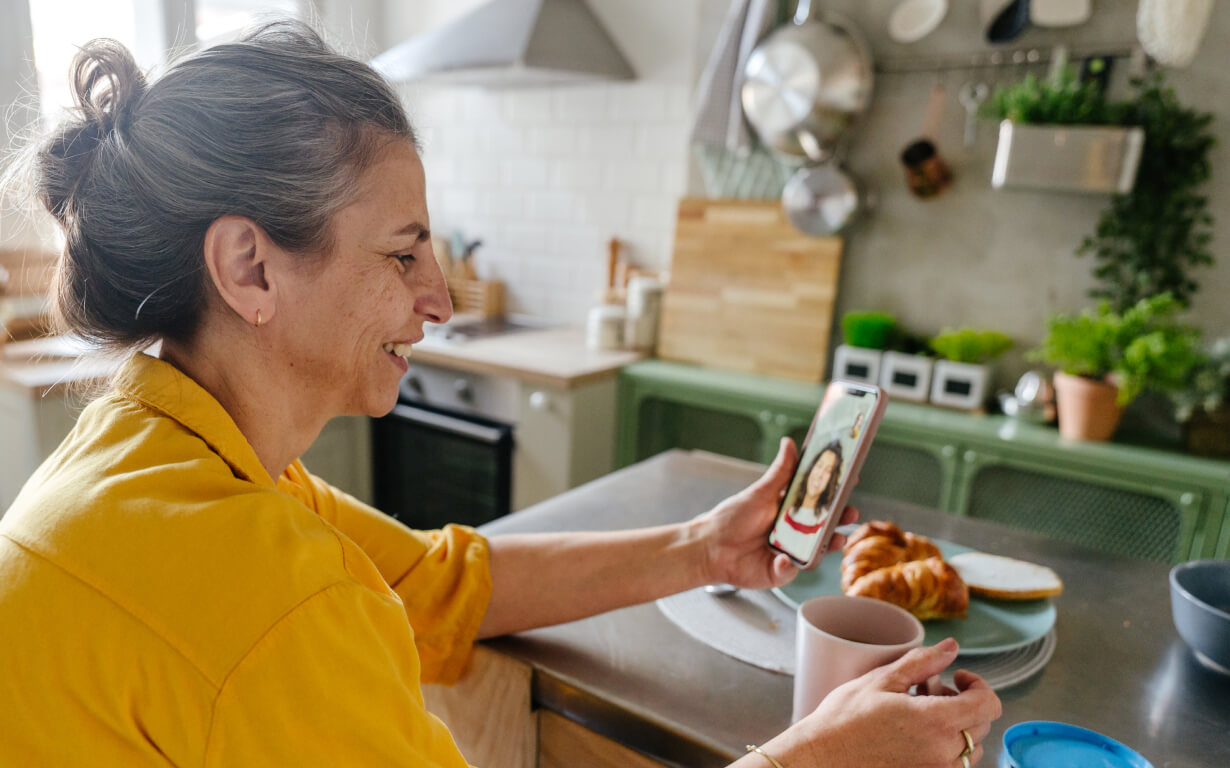 A woman in a yellow shirt is sitting at a kitchen counter, smiling at her phone during a video call. A croissant and toast are on a plate beside her, along with a cup. The kitchen has green cabinets and hanging pots.