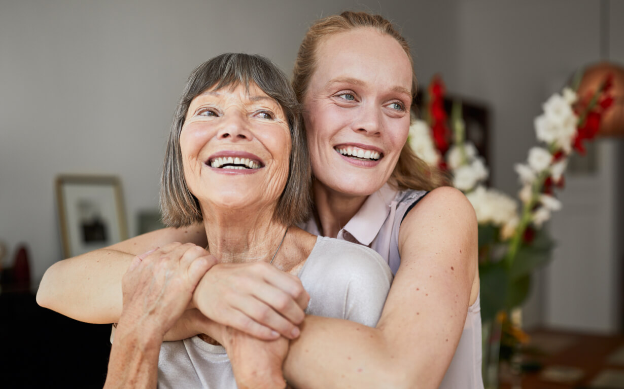 A smiling older woman and a younger woman from migrant communities embrace warmly in a well-lit room with flowers in the background. Both appear joyful and content, sharing a happy moment together.