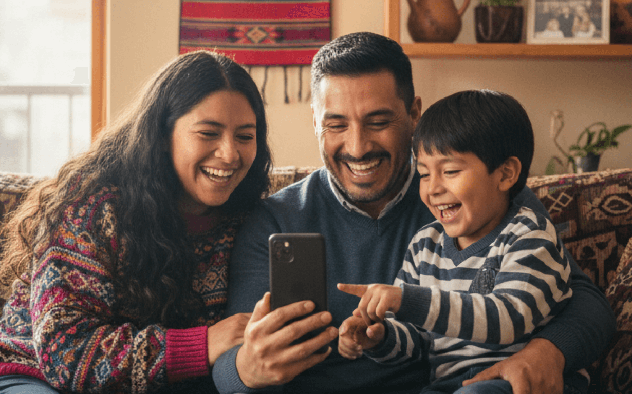 A smiling family of three, two adults and a child, sit on a couch looking at a smartphone together, laughing as they discover how easy it is to send money to Peru and share moments across the miles.
