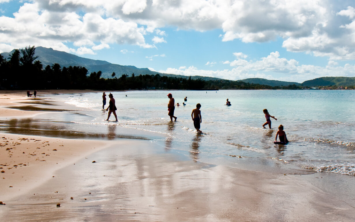 A scenic beach in Puerto Rico welcomes visitors with its tranquil ambiance, where people enjoy the pristine sandy shore under a partly cloudy sky. Hills and trees grace the distant shoreline, reflecting off the calm blue sea—a perfect snapshot for those considering moving to Puerto Rico serene beauty.
