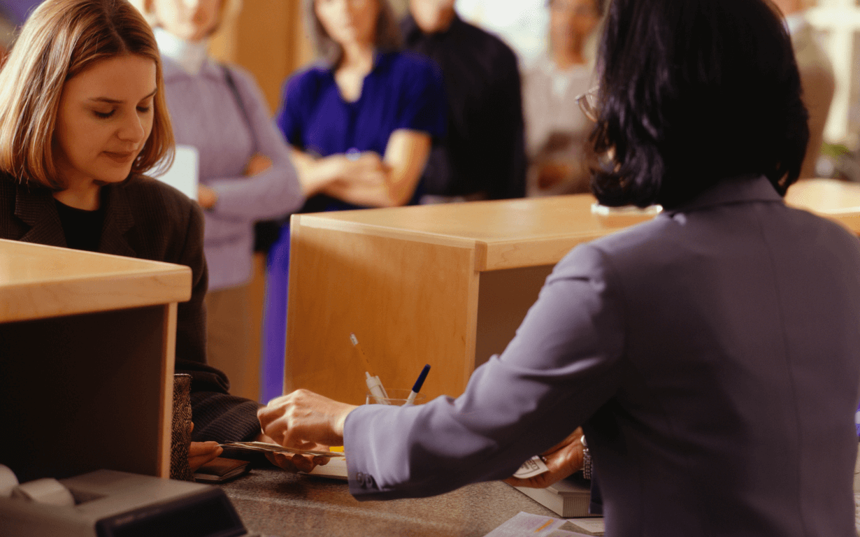 People at a bank, with one woman at the counter being assisted by a teller from one of the Best European Banks, while others wait in line behind her. The scene is busy, with focus on the interaction between the customer and the teller.