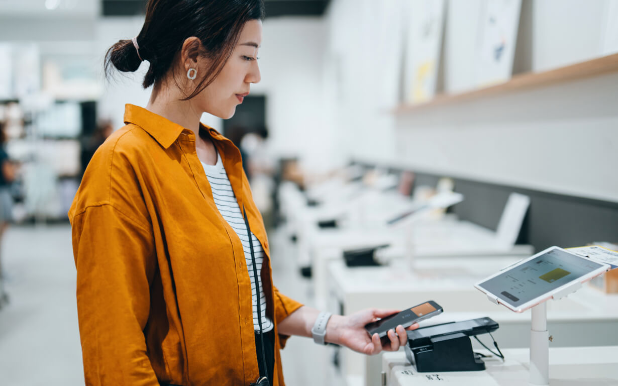 A woman in an orange jacket stands in a store, using her smartphone and digital wallet for a contactless payment at the terminal. She appears focused on the transaction, surrounded by various tech products displayed on the counters around her.