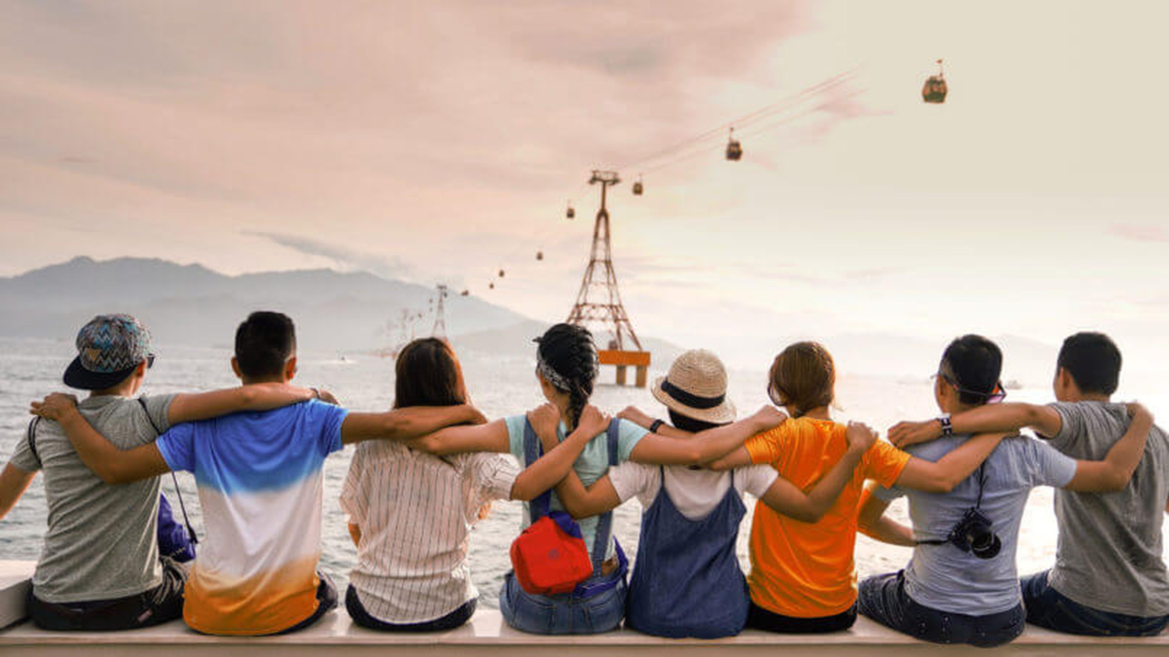 A group of seven people sit on a ledge with their backs to the camera, arms around each other, facing a scenic view of the water, mountains, and cable cars. It's sunset, the sky a blend of orange and pink hues. This heartwarming scene captures the essence of life and work post-COVID.