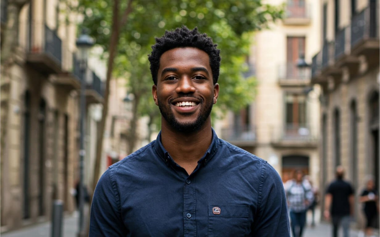 A man wearing a dark blue button-up shirt smiles while standing on a city street lined with trees and buildings, excited about moving to Spain, as people walk by in the background.