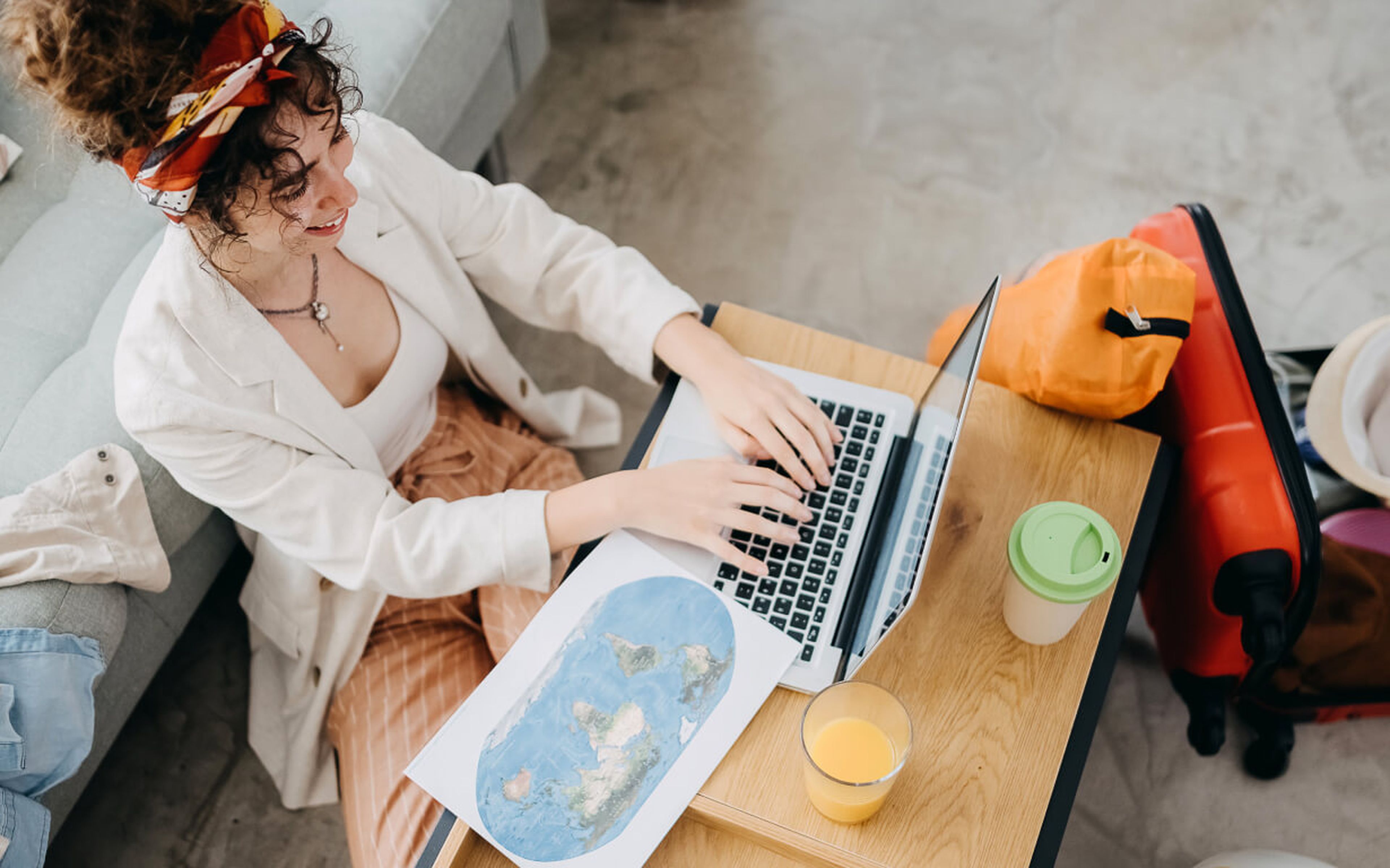 A person with curly hair tied in a headband is sitting on a sofa and typing on a laptop placed on a coffee table, preparing for a gap year. The table also has a map, a glass of orange juice, and a green cup. A red suitcase and an orange bag are on the floor nearby.