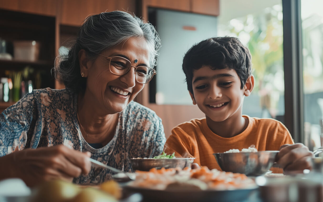 A smiling elderly woman and a young boy celebrate by enjoying a meal together at a table. The woman, in glasses and traditional attire, shares stories while the boy listens in his orange shirt, surrounded by shelves filled with cherished memories.