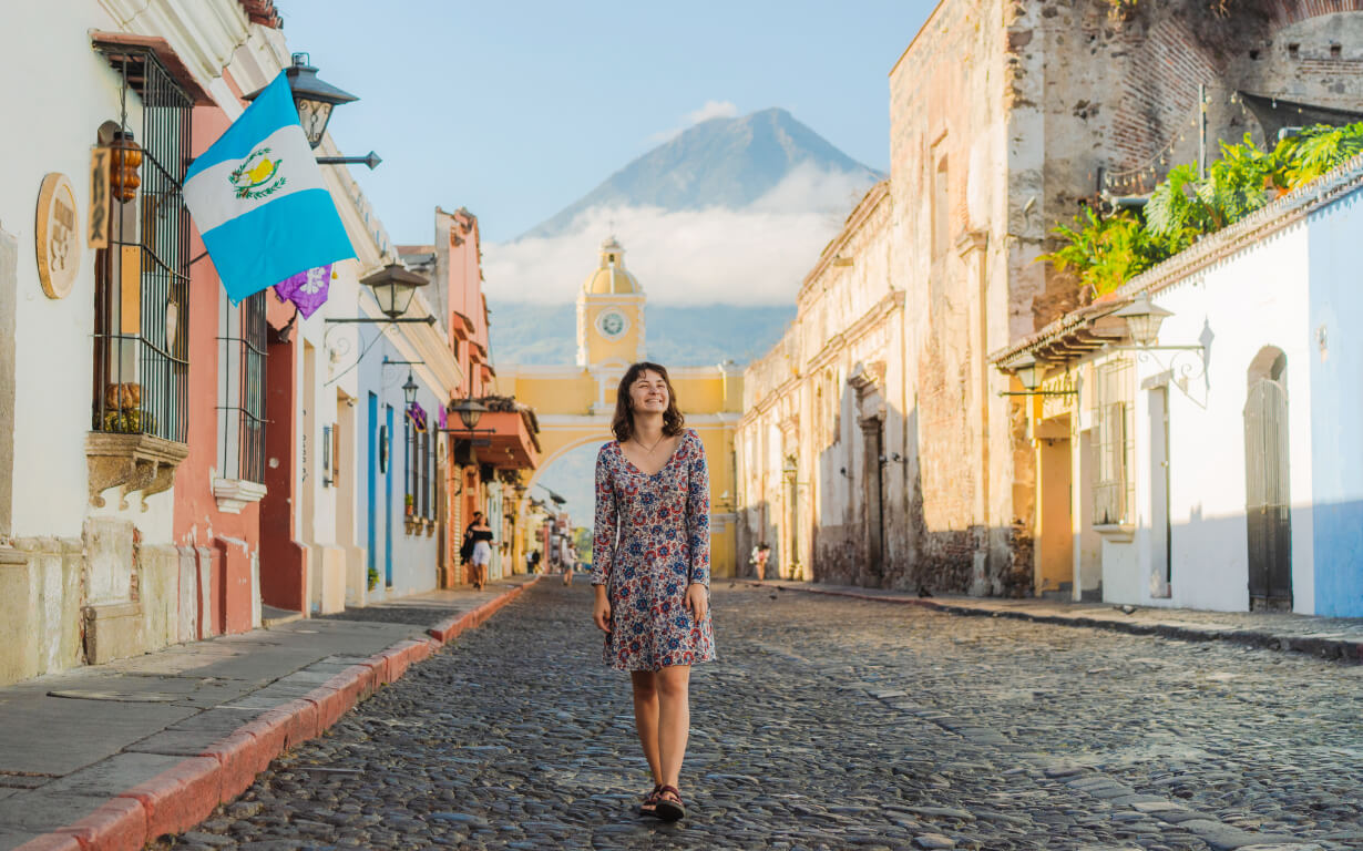 A woman in a floral dress walks down a cobblestone street in a historic town with colorful buildings. On the left, the Guatemalan flag waves proudly. In the background, a clock tower and a volcano capped with clouds loom over the scene.