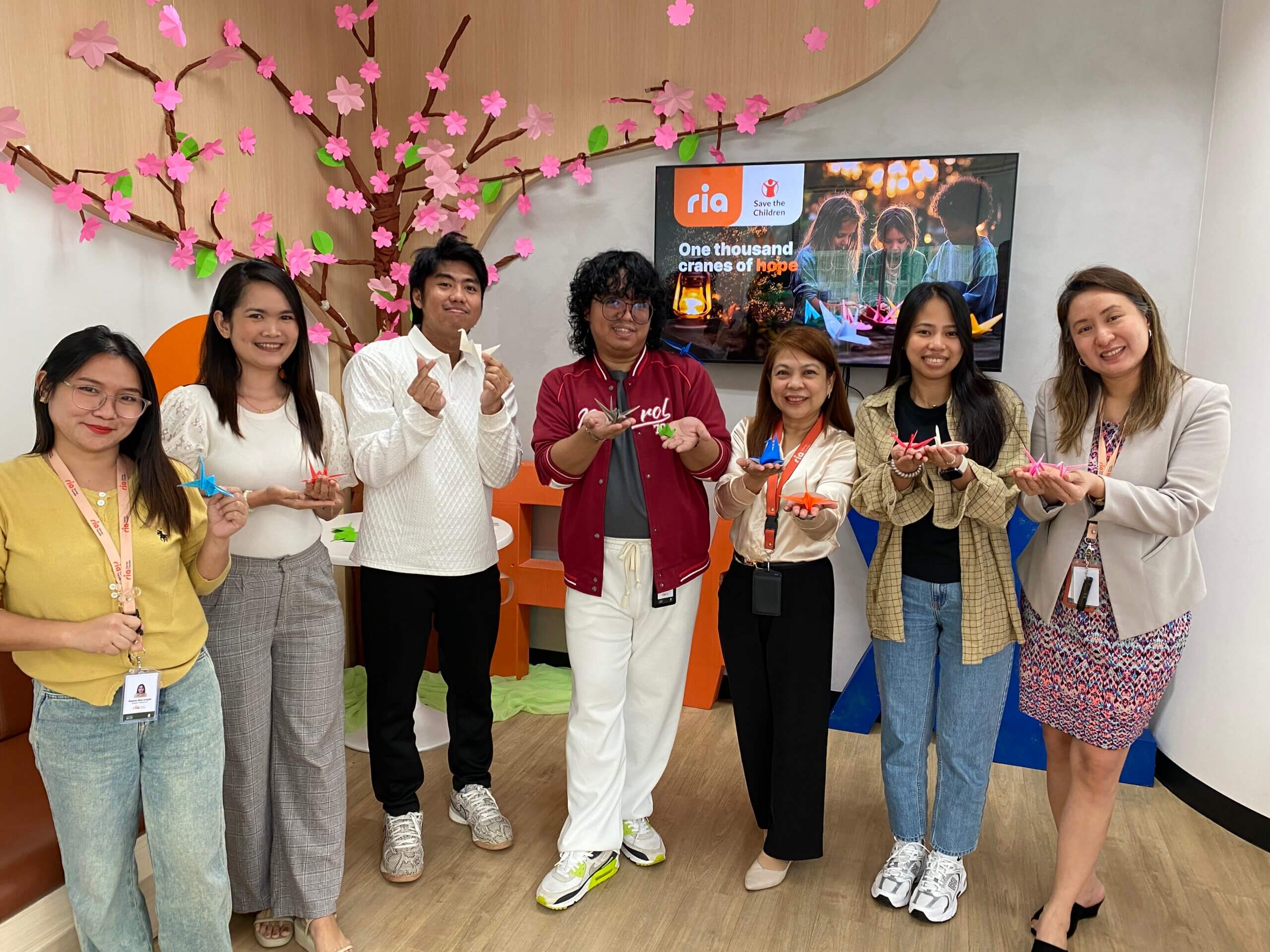 Seven people stand indoors, smiling and holding colorful origami cranes. They pose in front of a TV screen and a decorative pink paper tree. The setting appears casual and cheerful, likely an office or community space.