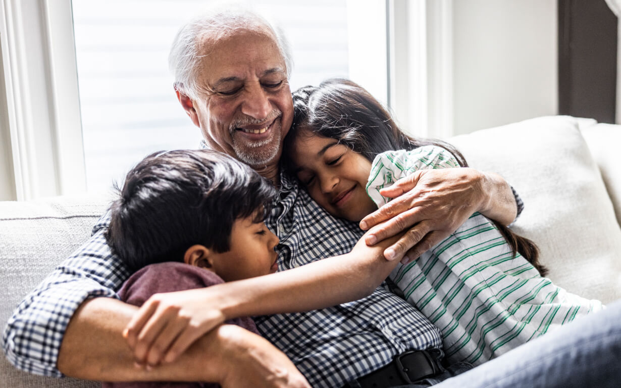 An elderly man with gray hair sits on a white couch, smiling warmly as he embraces two children, a boy and a girl, who are snuggling close to him with eyes closed. The room is softly lit by sunlight from large windows behind them.