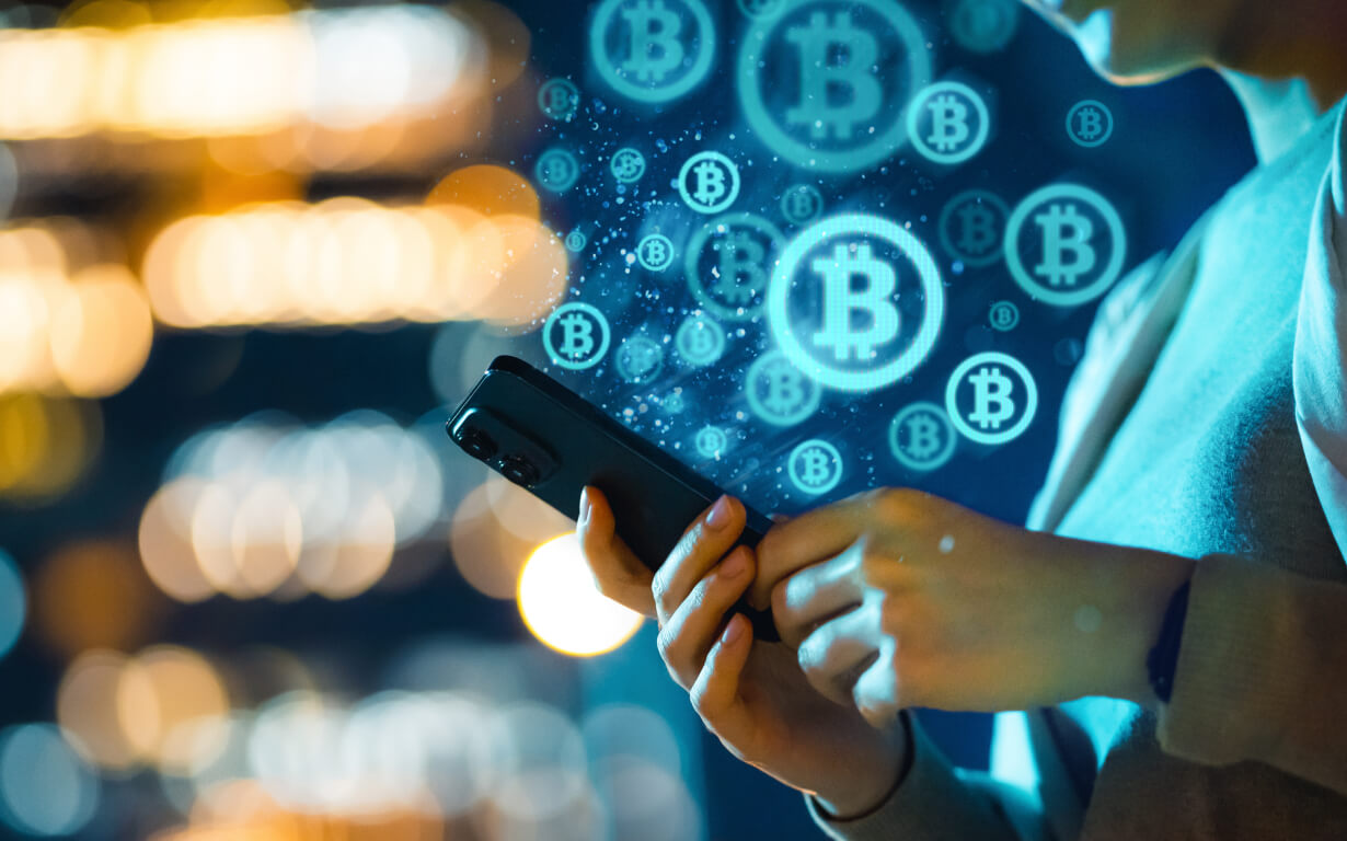 A person holding a smartphone with multiple glowing Bitcoin symbols floating in the air—one of the best ways to send money. The background features blurred lights, creating a sense of digital or financial technology.