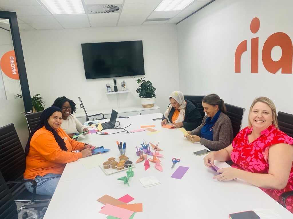 Five women sit around a conference table doing origami and smiling. Colorful paper, origami cranes, scissors, and muffins are on the table. A large screen and office decor are visible in the background.