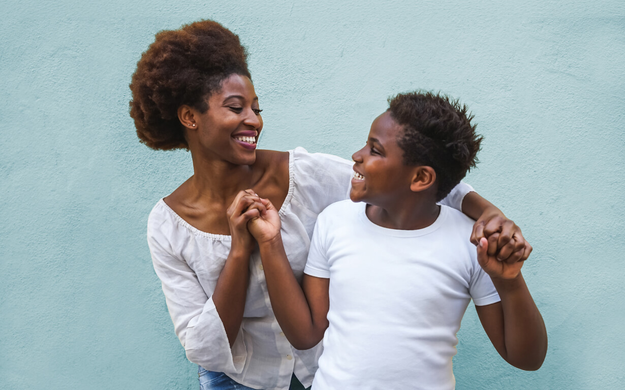 A joyful moment unfolds between a woman and a young boy, embodying the warmth often seen in migrant communities. They hold hands and smile at each other in front of a light blue wall. The woman is dressed in a white blouse, while the boy sports a white t-shirt.