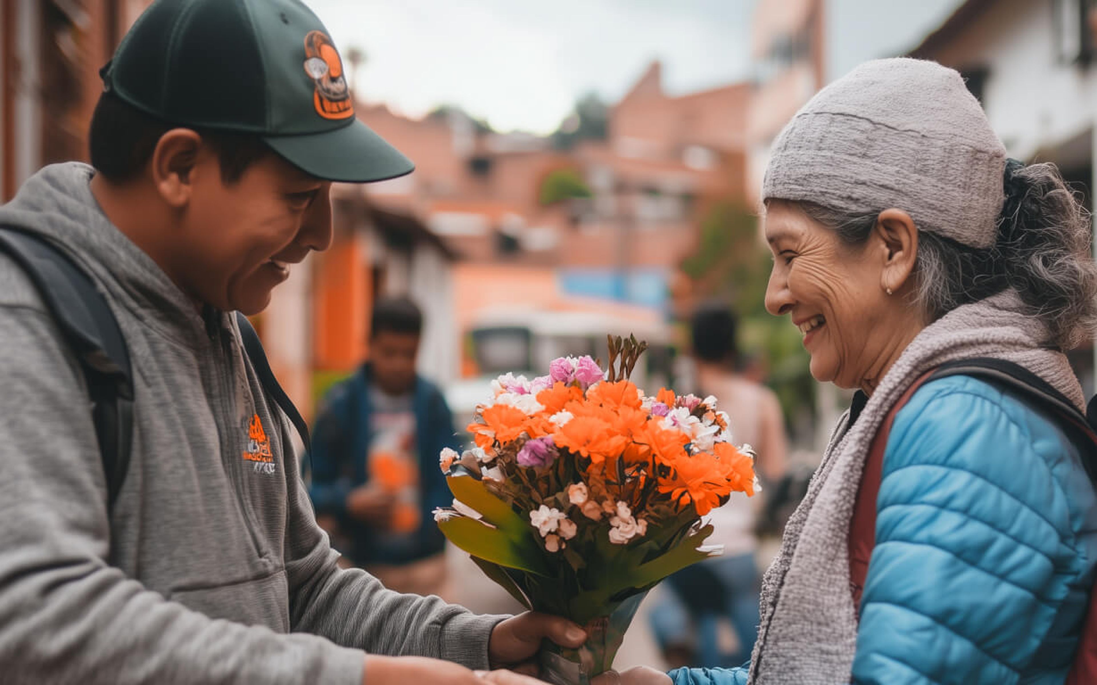 A smiling woman wearing a gray beanie receives a colorful bouquet from a man in a green cap, capturing the essence of how Mother's Day is celebrated. They stand outdoors on a street with blurred people and buildings in the background, radiating warmth and appreciation.