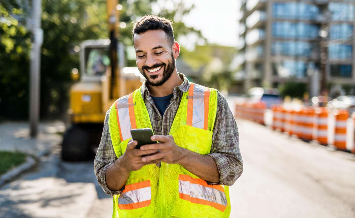 A smiling construction worker wearing a reflective safety vest checks his smartphone, possibly exploring how to send money without a bank account, while standing on a sunny street with construction equipment and orange traffic barriers in the background.
