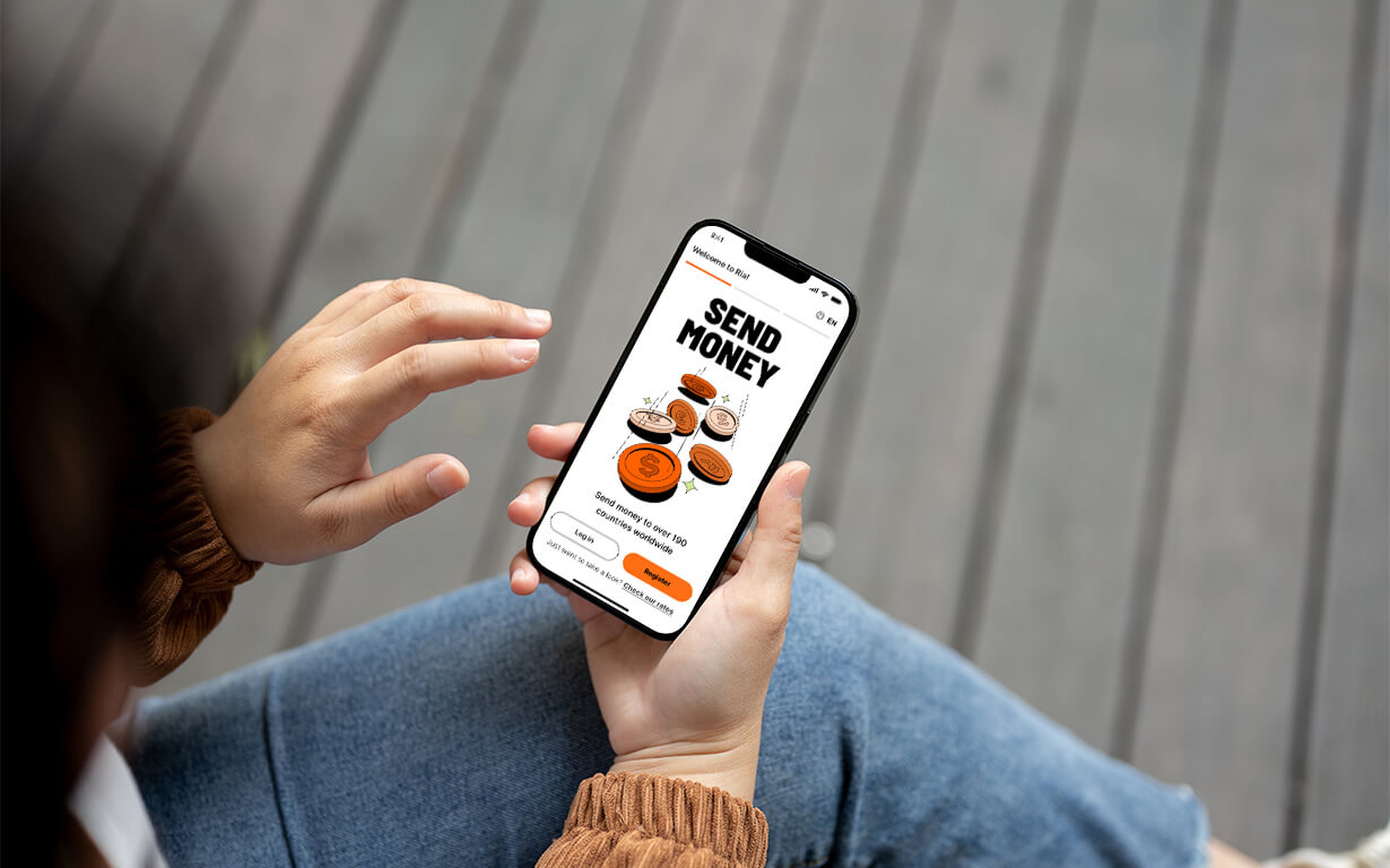 A person holding a smartphone on their lap uses the Ria app money transfer feature, with the text "Send Money" and an orange send button displayed on the screen.