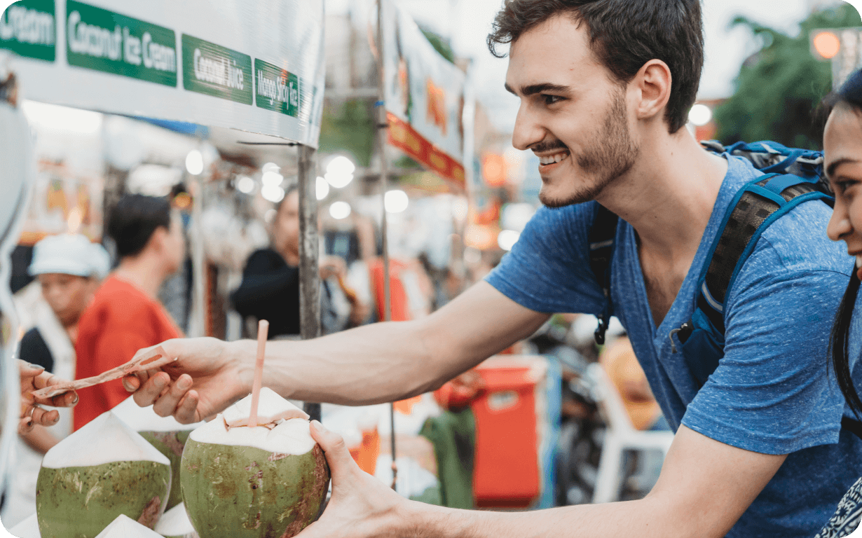A man in a blue shirt is smiling as he buys a fresh coconut at an outdoor market. He holds a coconut with a straw, surrounded by colorful food stalls and a vibrant, busy atmosphere.
