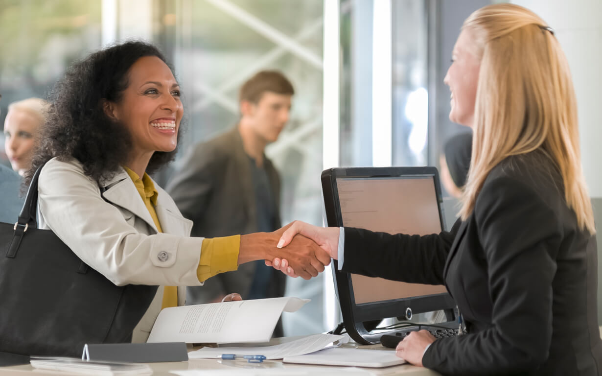 A woman in a gray coat and yellow blouse smiles as she shakes hands with a seated blonde woman in a black blazer across a desk adorned with a computer monitor displaying exchange rates. A blurred individual is visible in the background.