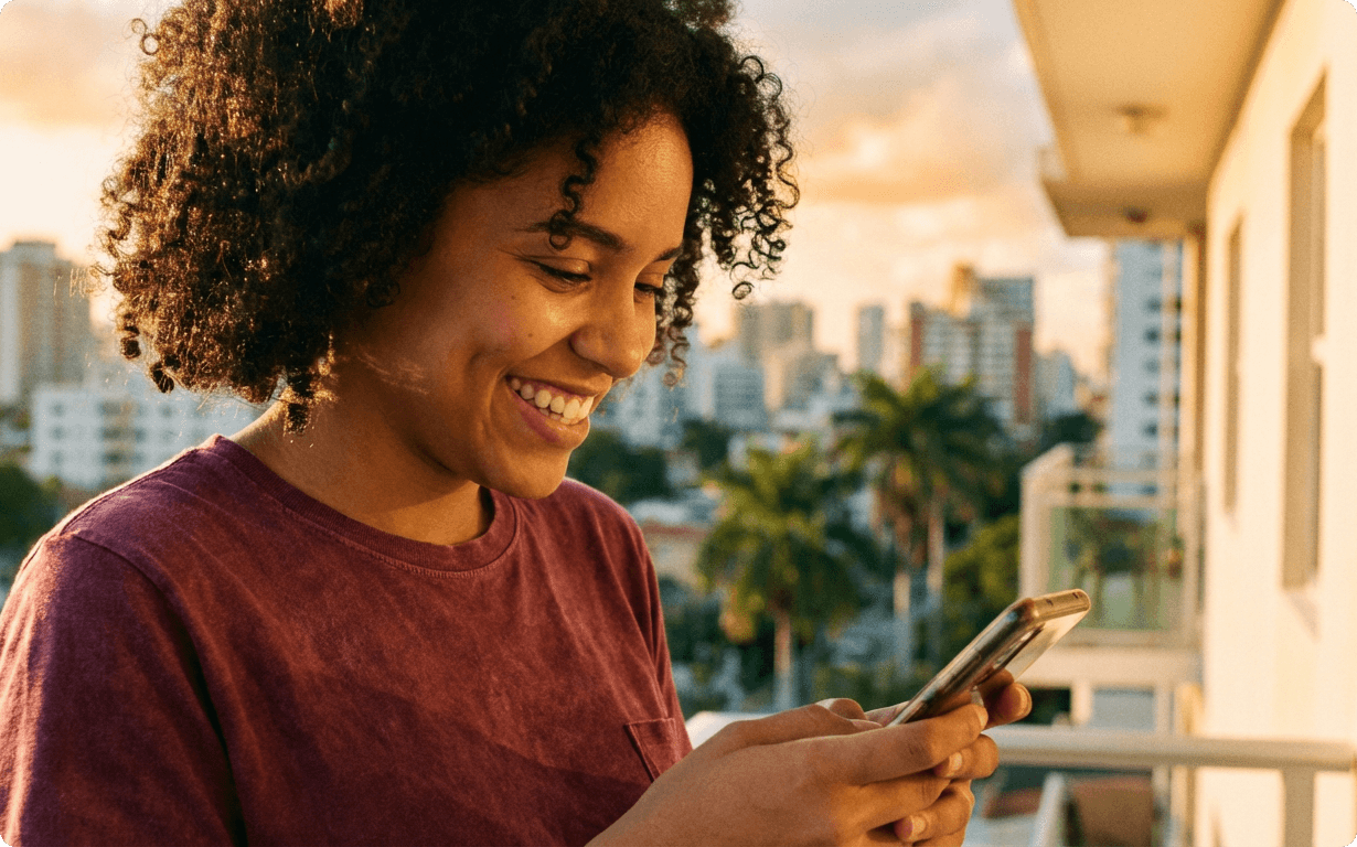 A young person with curly hair and a maroon shirt smiles while looking at a smartphone, standing outdoors at sunset with a city skyline and palm trees—perhaps getting ready to send money to Venezuela.