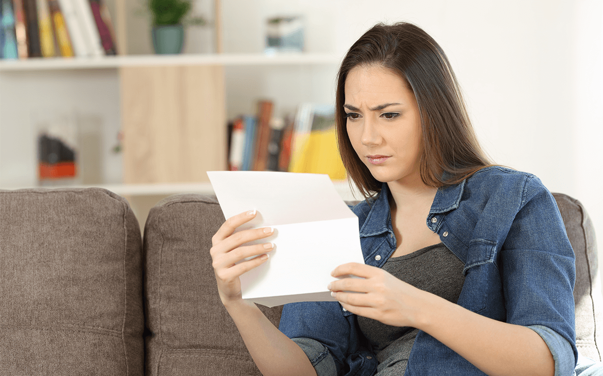 A woman with long brown hair, wearing a denim shirt, sits on a couch, intently examining a piece of paper that likely pertains to taxes. She appears puzzled or deep in concentration. In the background, a partially visible bookshelf hints at her interest in international money transfers.