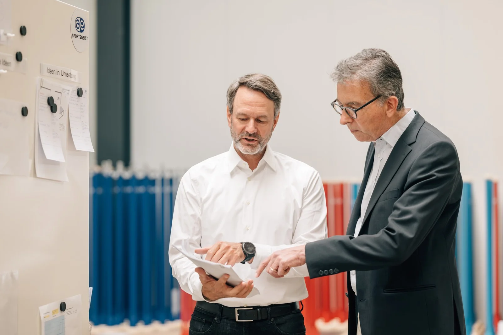 Zwei Männer besprechen Unterlagen an einer Pinnwand in einer Produktionshalle