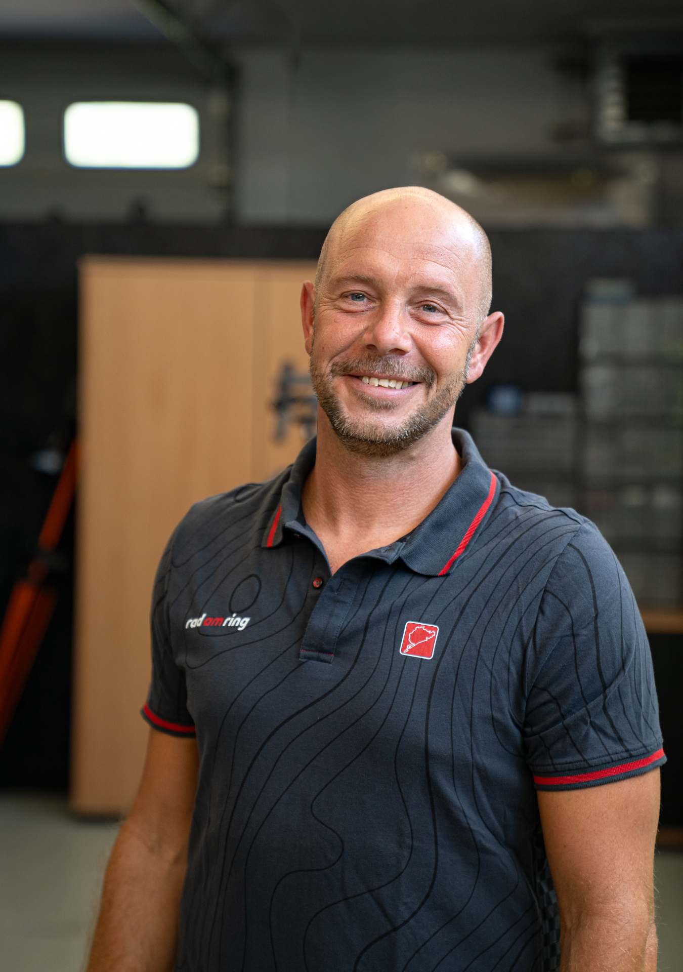 Ein lächelnder Mann mit Glatze und Bart in einem dunkelgrauen Rad am Ring Poloshirt mit Nürburgring-Logo.