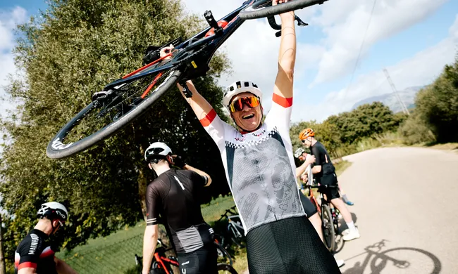 Glücklicher Radfahrer in Trikot hält sein Rennrad über dem Kopf, jubelnd mit anderen Radfahrern im Hintergrund.