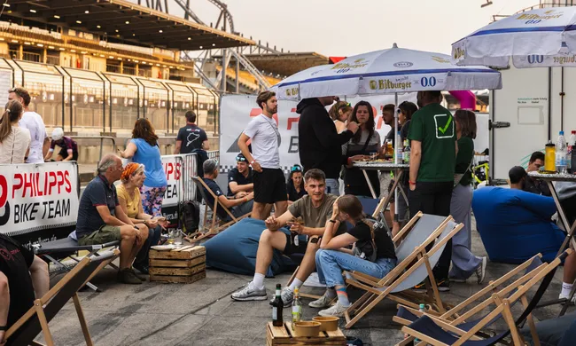 Menschen entspannen im Freien auf Liegestühlen und Sitzsäcken. Im Hintergrund sind Tribünen und ein "Philipps Bike Team" Banner zu sehen. Bitburger Schirme spenden Schatten.