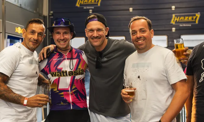 Vier Männer lächeln in die Kamera, umarmen sich und halten Biergläser. Einer trägt ein buntes Radtrikot, im Hintergrund "HAZET"-Logo.