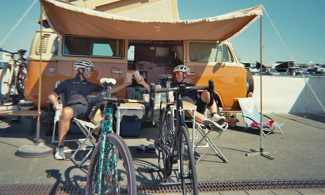 Zwei Radfahrer entspannen sich nach der Fahrt vor einem orangefarbenen VW Bus mit Markise. Rennräder stehen im Vordergrund.