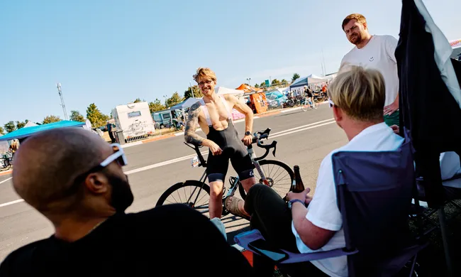 Lächelnder Radfahrer in Sportkleidung lehnt an seinem Rad und spricht mit Freunden auf einem sonnigen Outdoor-Event mit Zelten im Hintergrund.