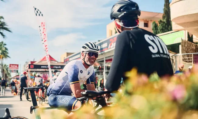 Ein lächelnder Radfahrer mit Helm und Sonnenbrille unterhält sich bei einem sonnigen Outdoor-Event mit einem anderen Radfahrer.