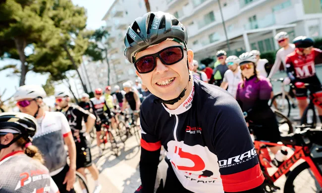 Ein lächelnder Radfahrer in Helm, Sonnenbrille und Trikot blickt in die Kamera, umgeben von anderen Radsportlern auf einer sonnigen Straße.