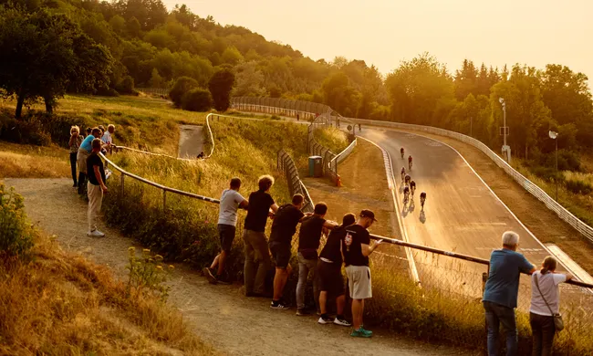 Zuschauer beobachten Radfahrer auf einer kurvigen Rennstrecke bei Sonnenuntergang in einer hügeligen, grünen Landschaft.