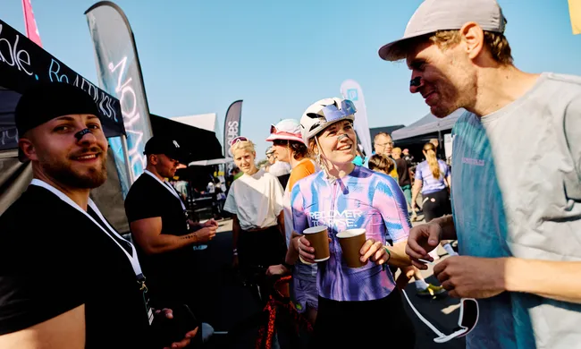 Lachende Radsportler und Besucher mit Nasenpflastern unter blauem Himmel bei einem Outdoor-Event, unterhalten sich und halten Becher.