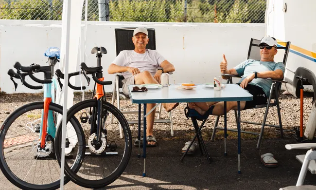 Zwei Männer entspannen sich an einem Tisch im Freien, neben zwei Rennrädern und einem Wohnmobil. Einer gibt einen Daumen hoch.