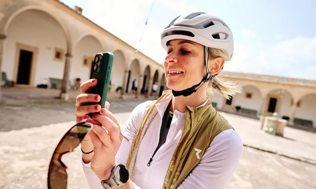 Lächelnde Frau mit Fahrradhelm und Weste fotografiert mit Smartphone in einem sonnigen Innenhof mit Arkaden.