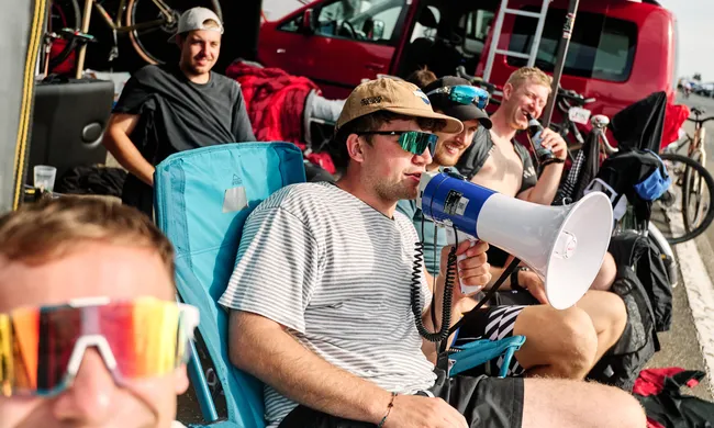 Junge Männer entspannen in Campingstühlen vor einem roten Van, einer hält ein Megafon, ein anderer trinkt. Im Vordergrund unscharfes Gesicht mit Sonnenbrille.