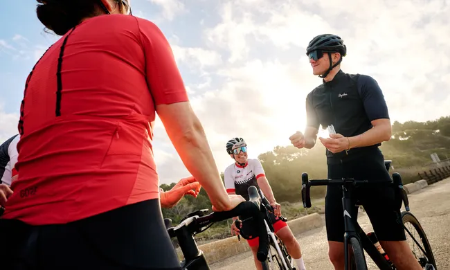 Drei Radfahrer unterhalten sich bei einer Pause im warmen Abendlicht. Ein Mann isst einen Snack, eine Frau lächelt.