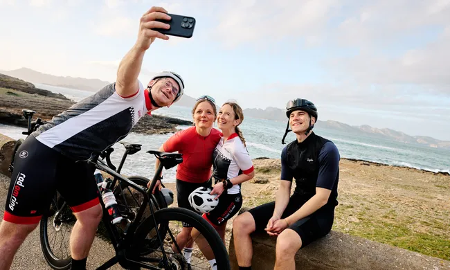 Vier fröhliche Radfahrer machen ein Selfie an einer felsigen Küste, mit Meer und Bergen im Hintergrund.