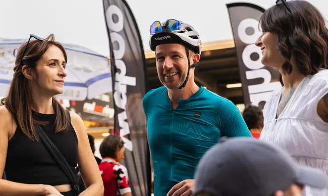 Drei Personen, darunter ein lächelnder Radfahrer im türkisen Trikot, unterhalten sich angeregt bei einem Outdoor-Event.